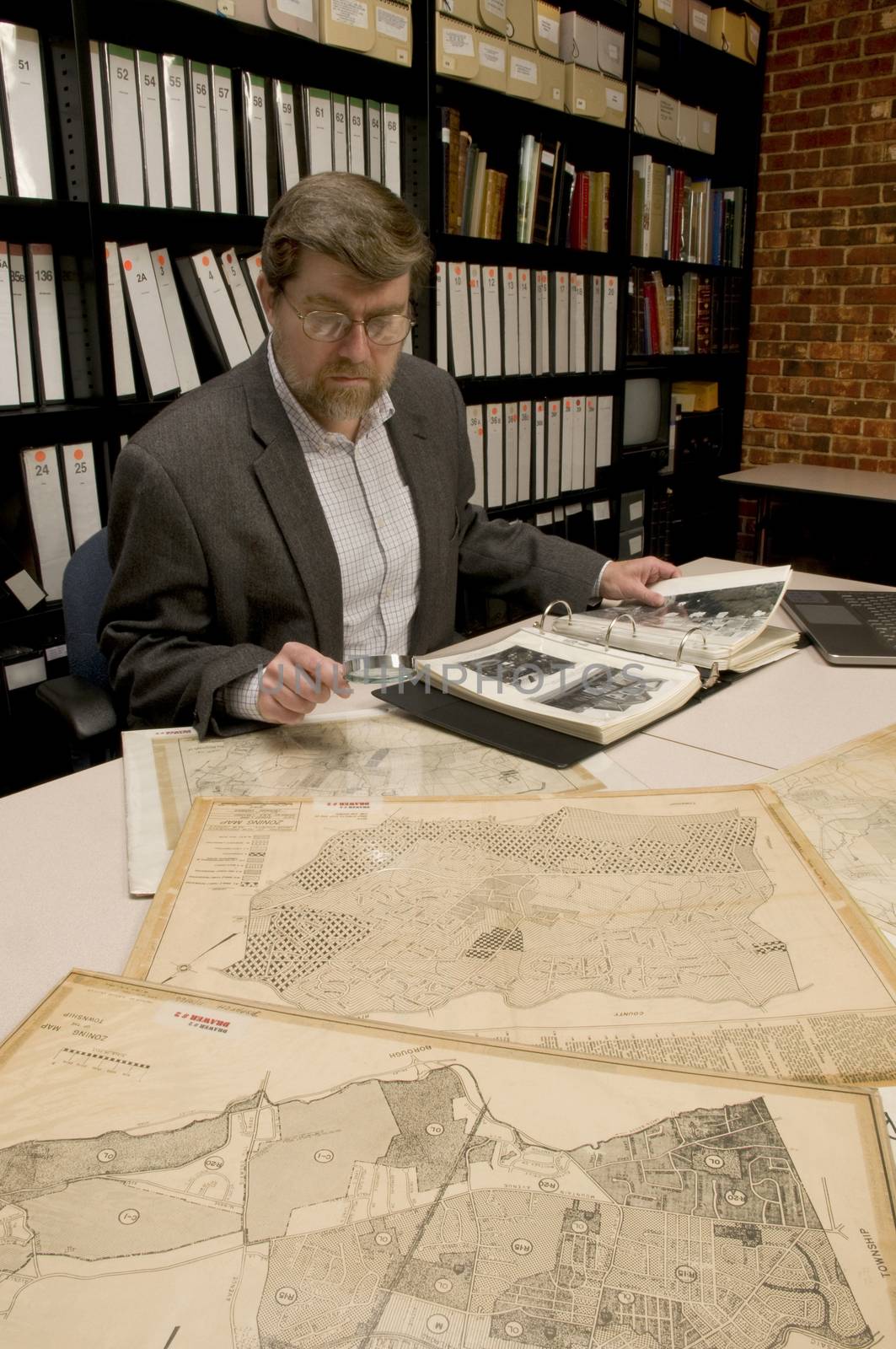 Researcher in archive, searching through maps and photographs. [Maps and photographs on table are public domain. Image on computer creted for this photograph. Model and Property Released.]