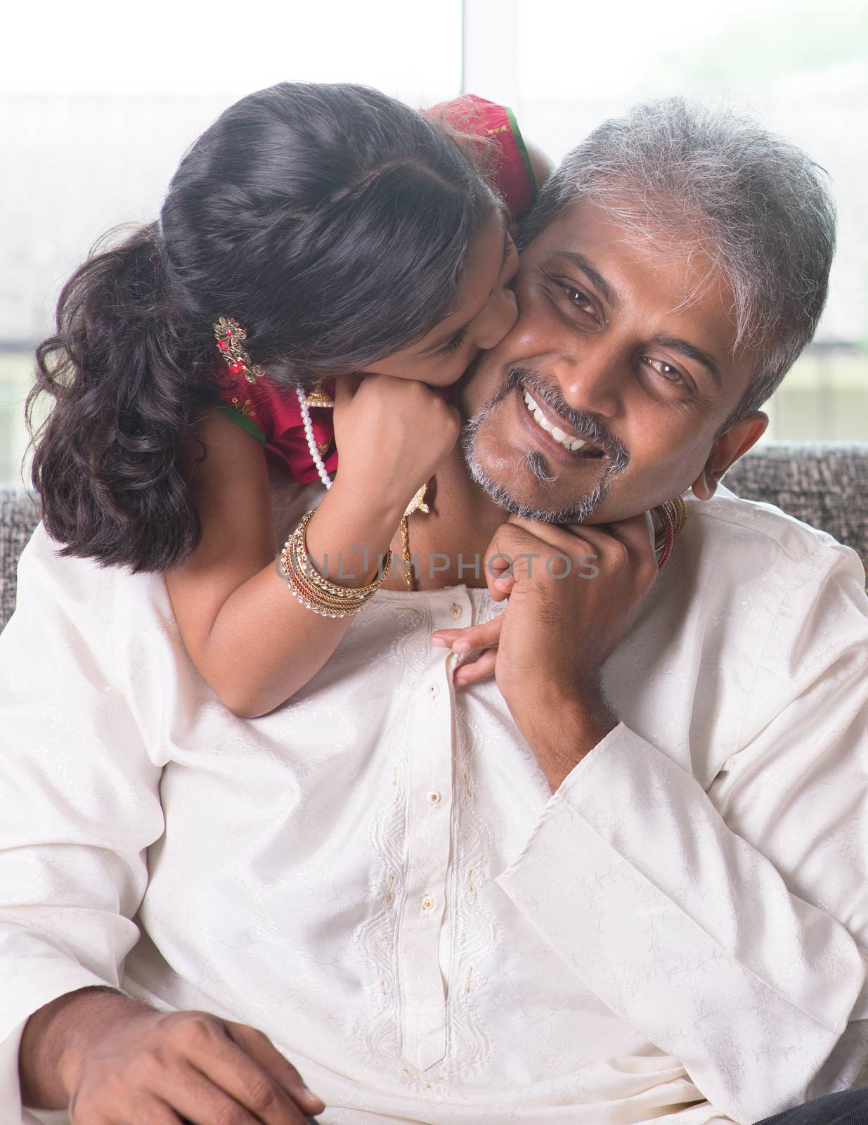 Happy Indian family at home. Asian girl kissing her father, sitting on sofa. Parent and child indoor lifestyle.