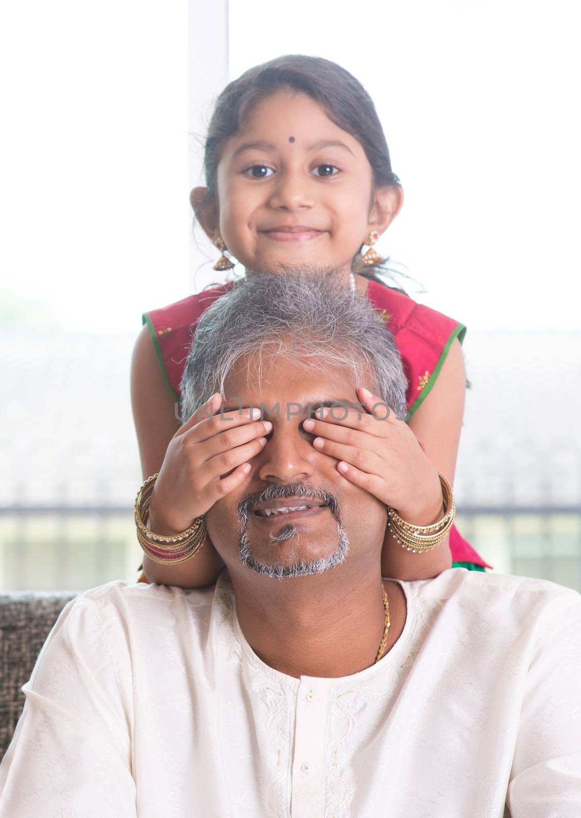 Happy Indian family at home. Asian girl giving surprise to her dad by covering dad eyes. Father and daughter indoor lifestyle.