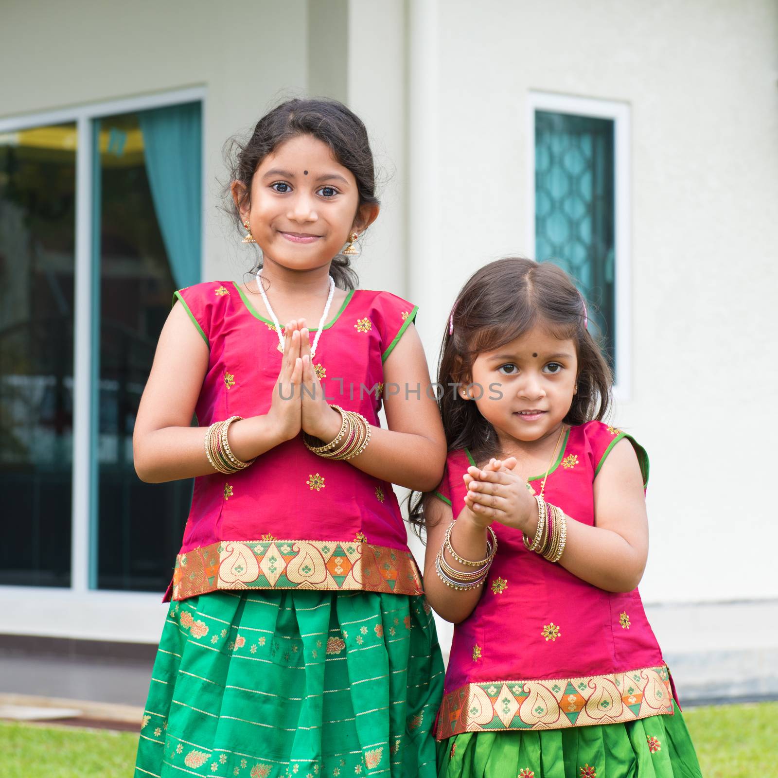 Cute Indian girls dressed in sari with folded hands representing traditional Indian greeting, standing outside their new house celebrating diwali, festival of lights.
