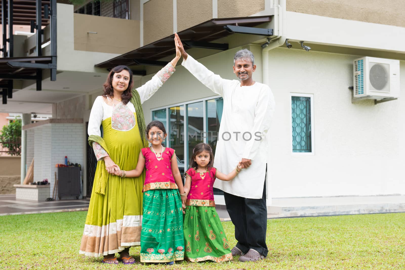 Parents forming house roof shape above children. Beautiful Asian Indian family portrait smiling and standing outside their new house.