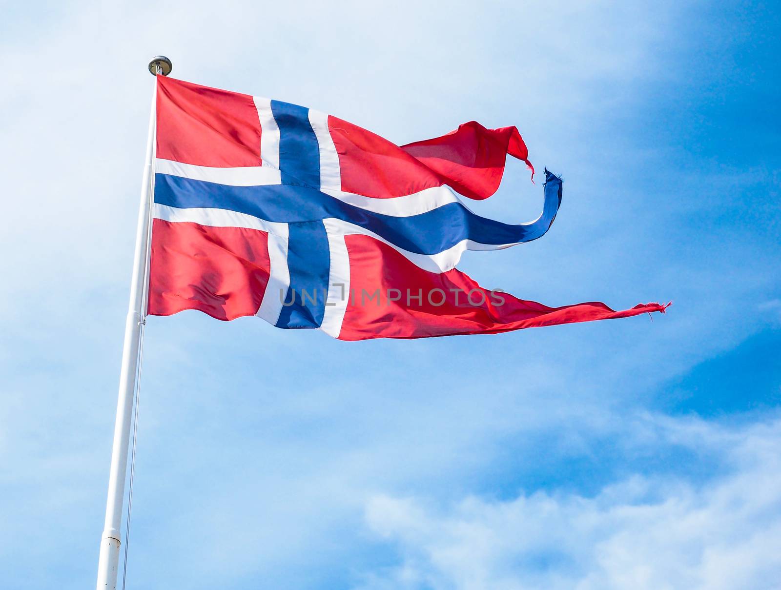 The Royal flag of Norway on a pole towards blue and white sky by Arvebettum