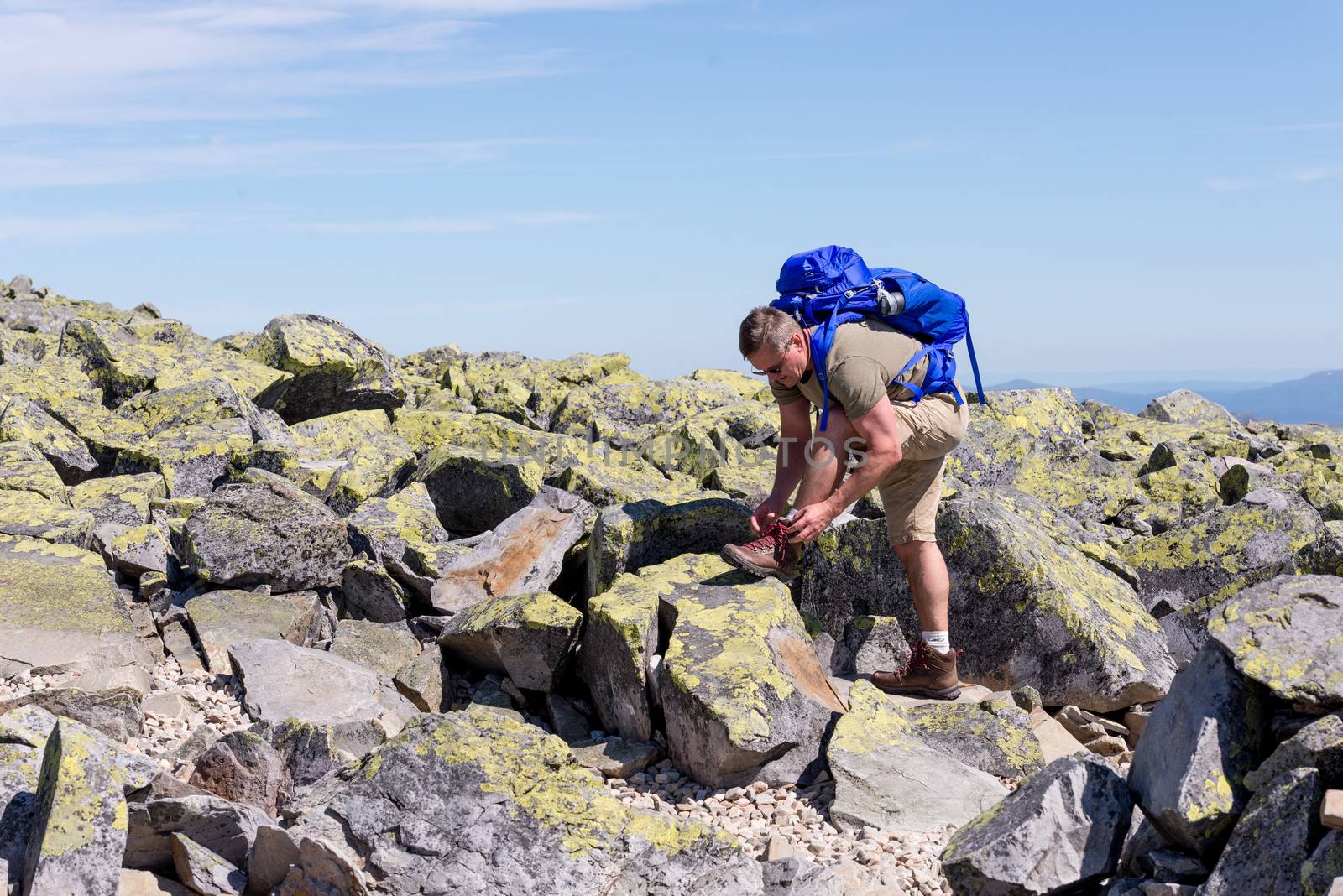 Hiker with big traveling rucksack tying up laces on the mountain trail, adventure travel and discovery