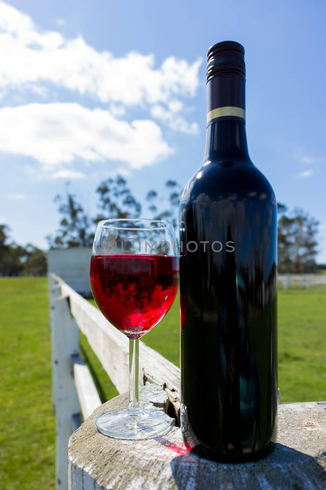 A red wine bottle and glass resting on a white fence in the country.