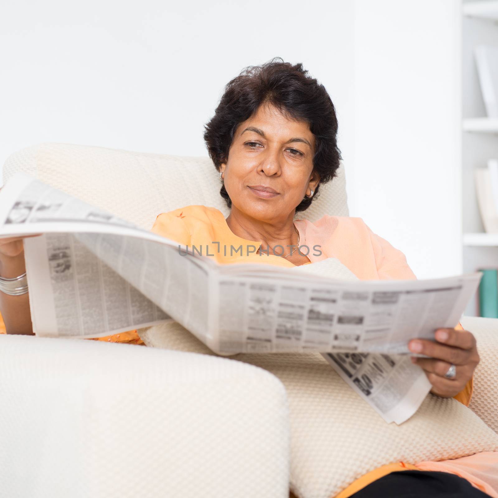 Portrait of a 50s Indian mature woman reading newspaper, sitting on sofa at home. Indoor senior people living lifestyle.