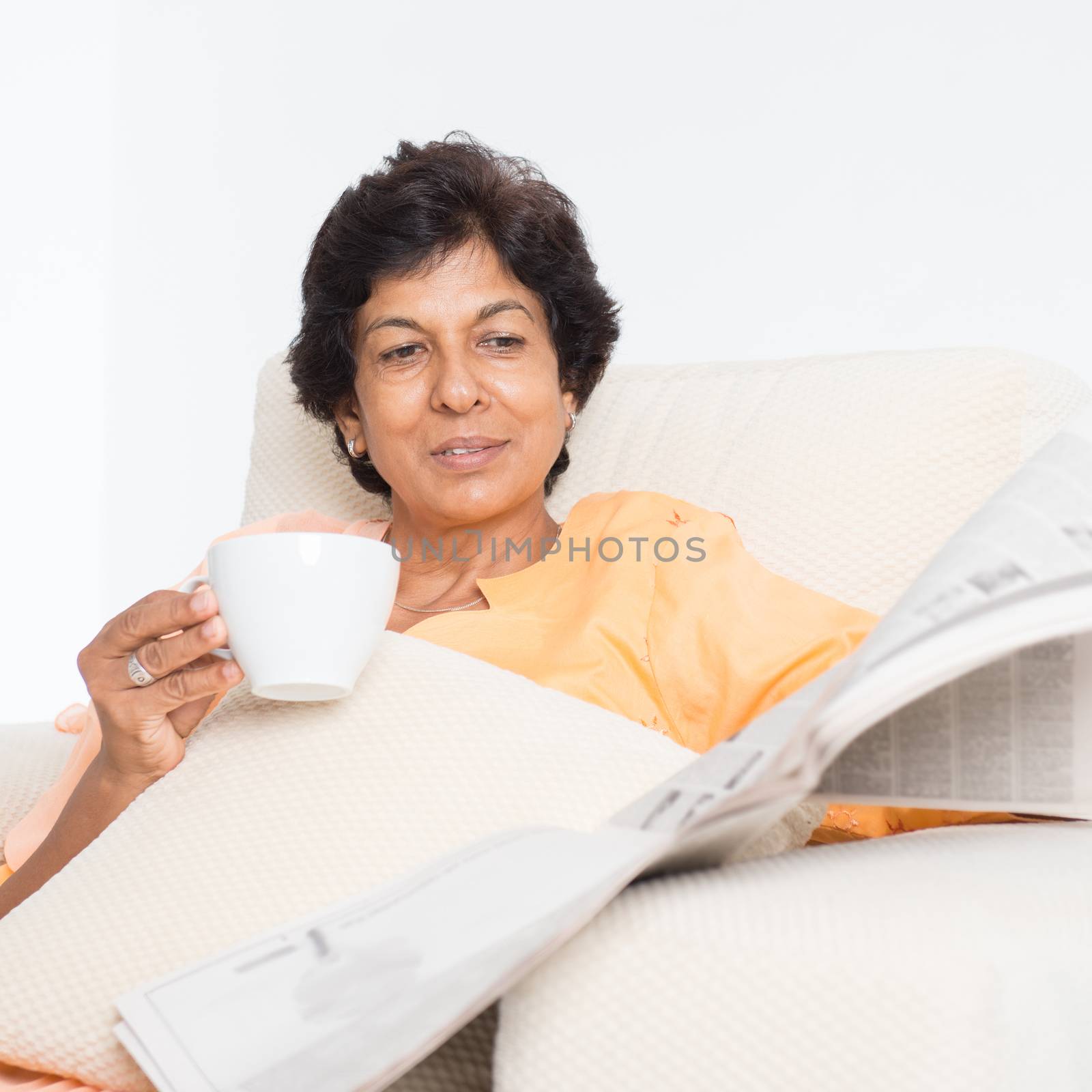 Portrait of a 50s Indian mature woman reading newspaper and drinking coffee at home. Indoor senior people living lifestyle.