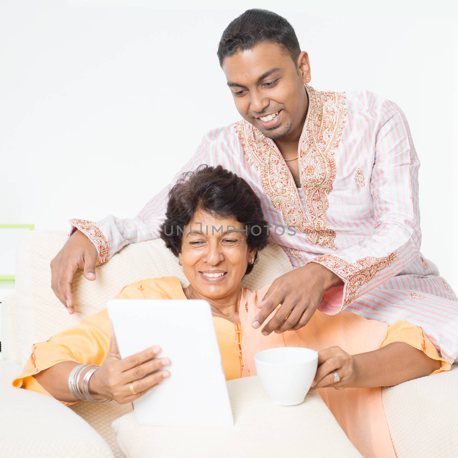 Portrait of happy Indian family using mobile apps with tablet pc at home. Beautiful mature 50s Indian mother and her 30s grown son.