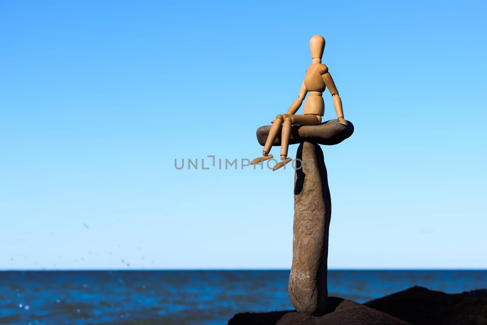 Wooden mannequin sitting on the top of sea stone