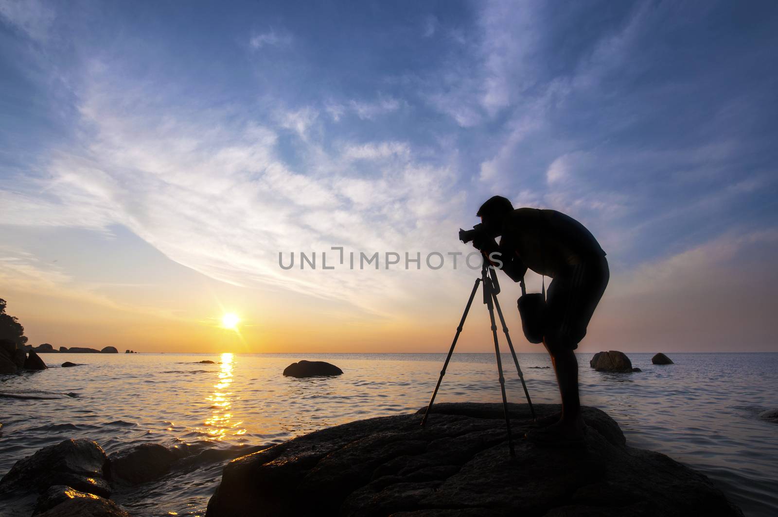 Silhouette a photographer taking pictures of sunrise on a rock, near the tropical beach.
