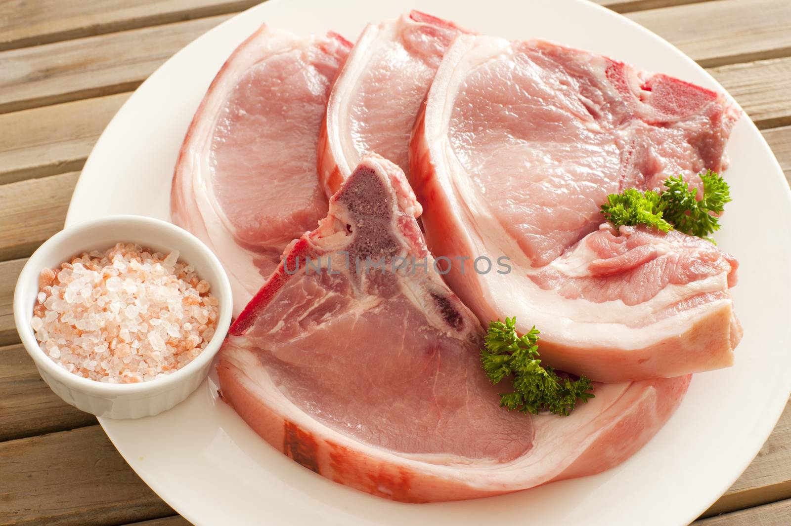 Close up Fresh Pork Chops with Herbs on a White Round Plate with Rock Salt on a Saucer.
