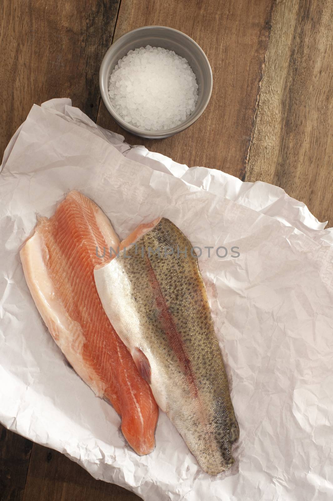Top View of Fresh Rainbow Trout Meat on a Paper Placed on Wooden Table with Rock Salt on a Saucer.