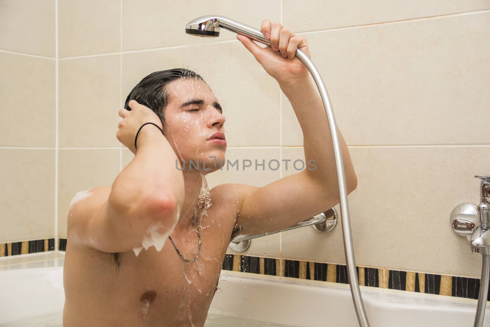 Handsome young man in bathtub at home having bath, washing body and hair with bathfoam and shampoo