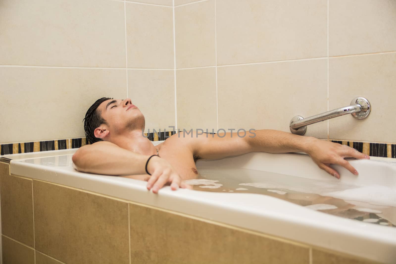 Handsome young man in bathtub at home having bath, washing body and hair with bathfoam and shampoo