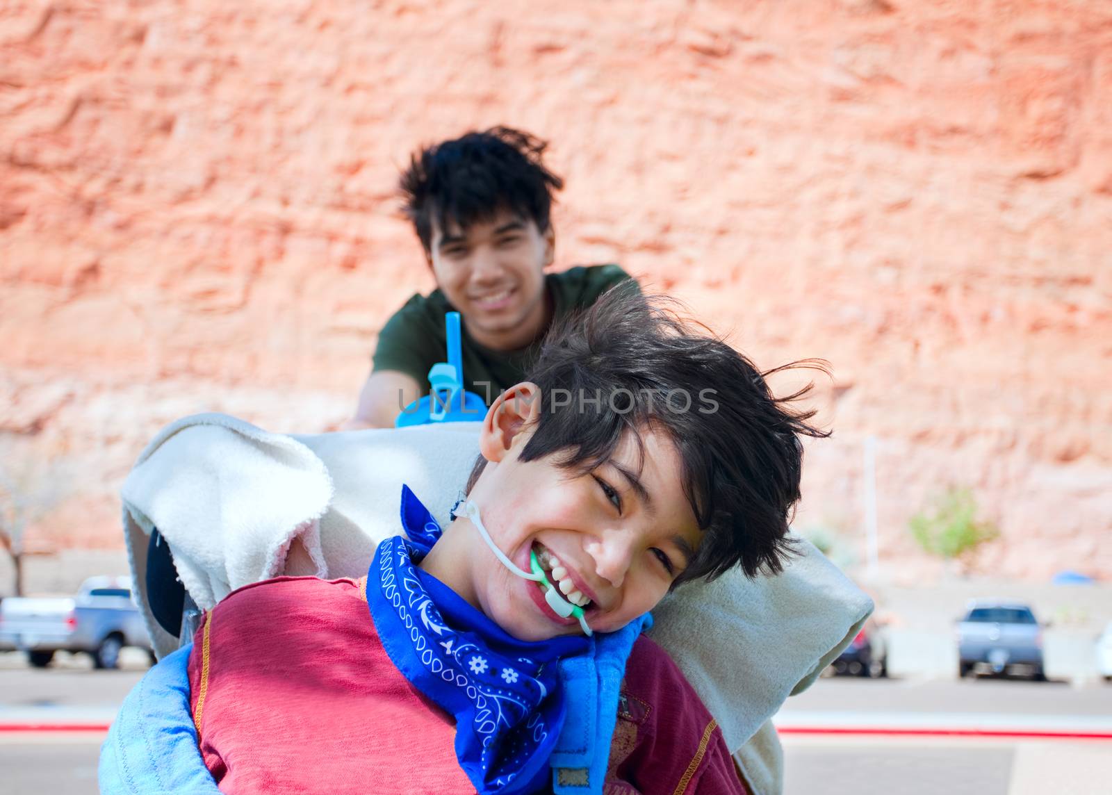 Teen boy pushing happy disabled biracial little  brother in wheelchair, smiling and laughing, outdoors. cerebral palsy