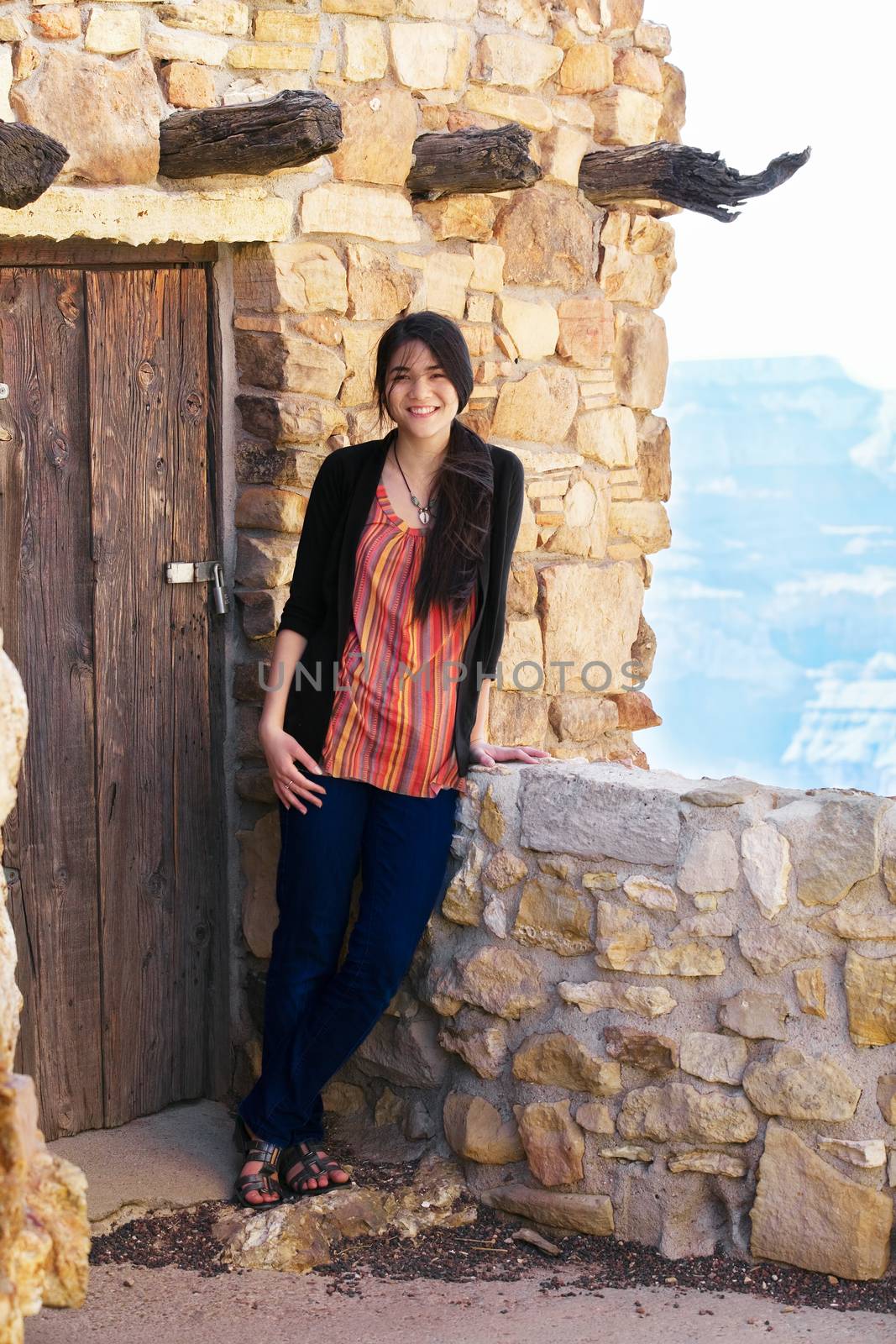 Biracial teen girl relaxing, leaning against rock wall overlooking Grand Canyon, next to doorway