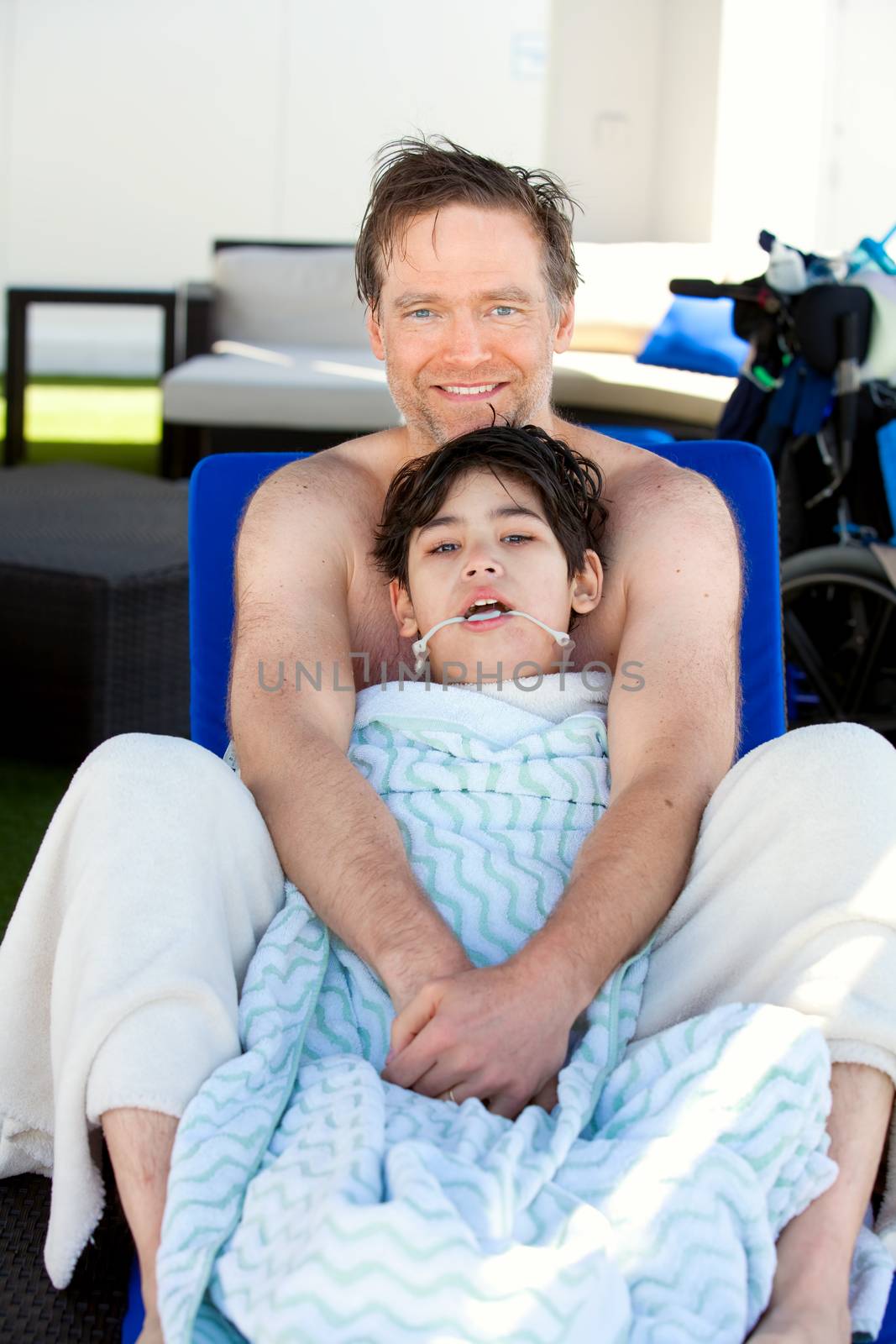 Caucasian father holding disabled son on blue poolside lounger, drying off after swimming. Child has cerebral palsy. 