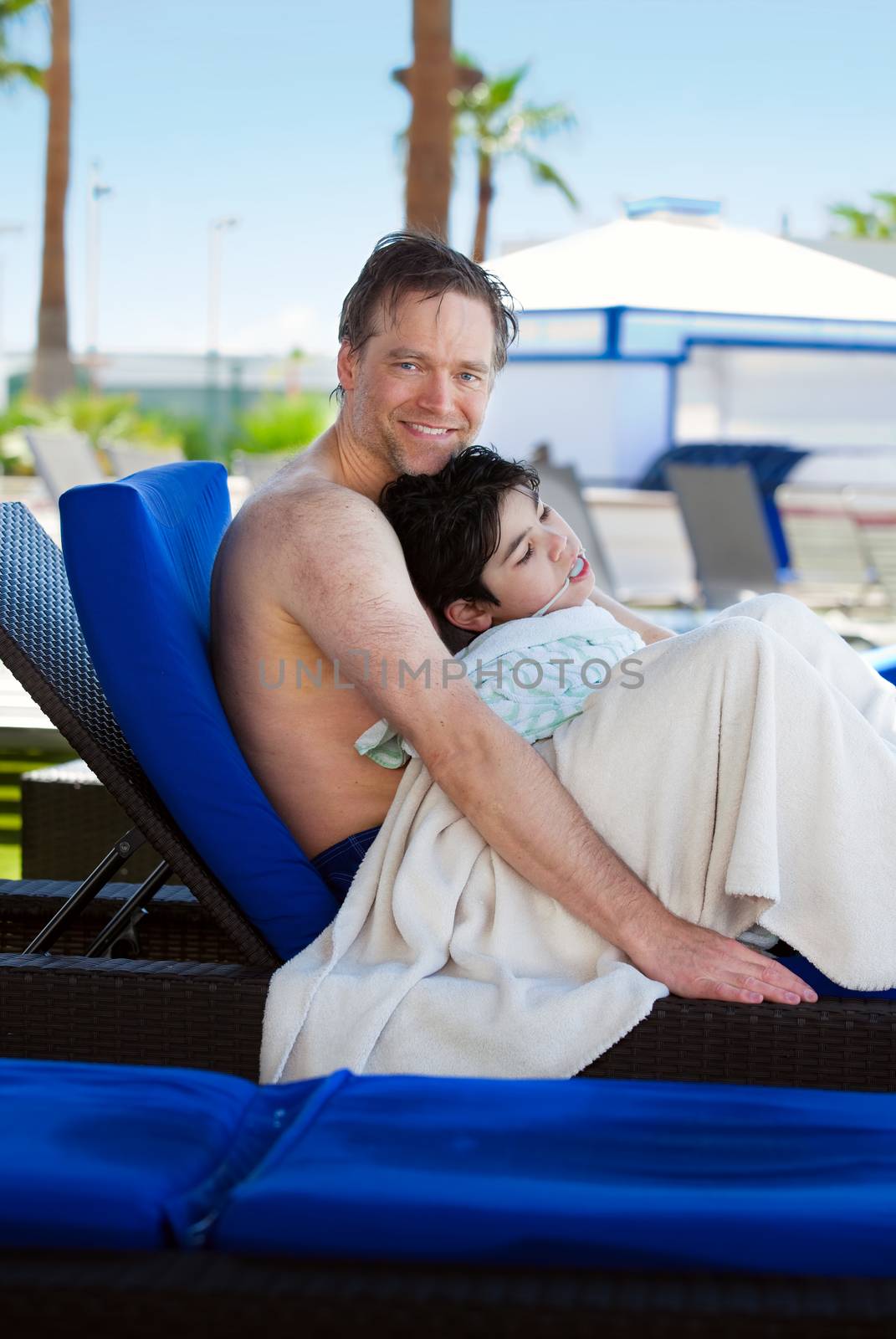 Caucasian father holding disabled son on blue poolside lounger, drying off after swimming. Child has cerebral palsy. 