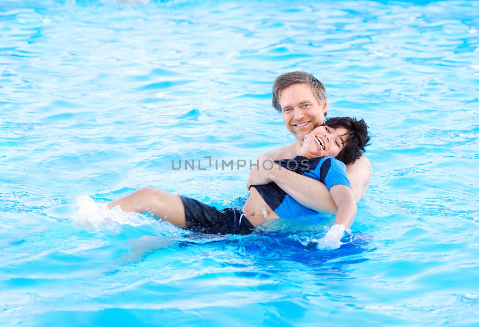 Caucasian father swimming in pool with biracial disabled son in his arms. Child has cerebral palsy.