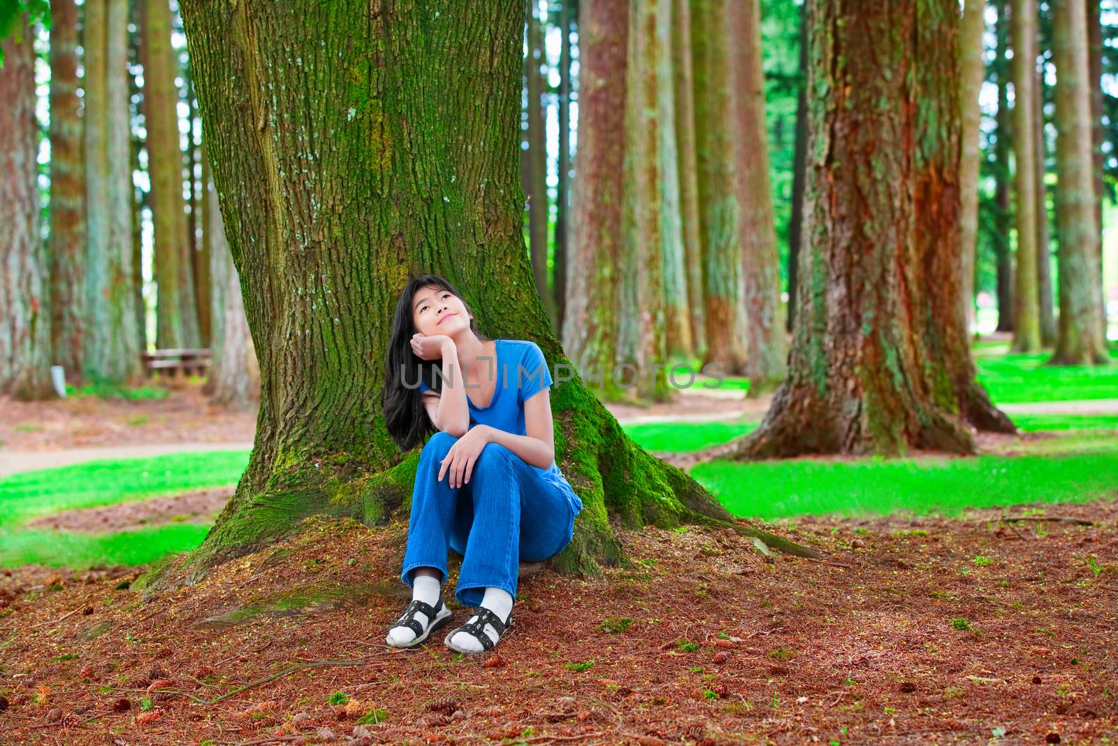 Young teen girl sitting under large pine trees, thinking by jarenwicklund