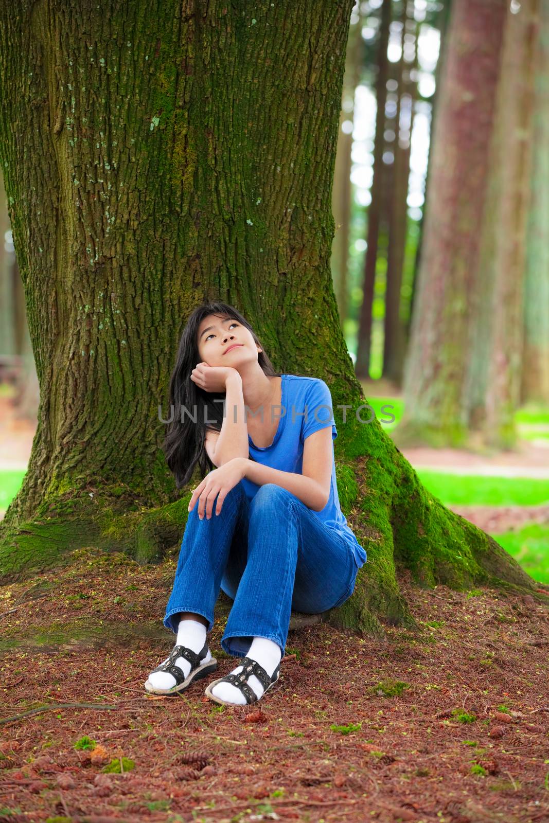 Young teen girl sitting under large pine trees, thinking by jarenwicklund