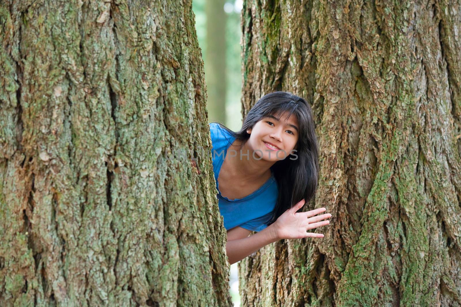 Biracial young teen girl peeking between trees, waving and smiling