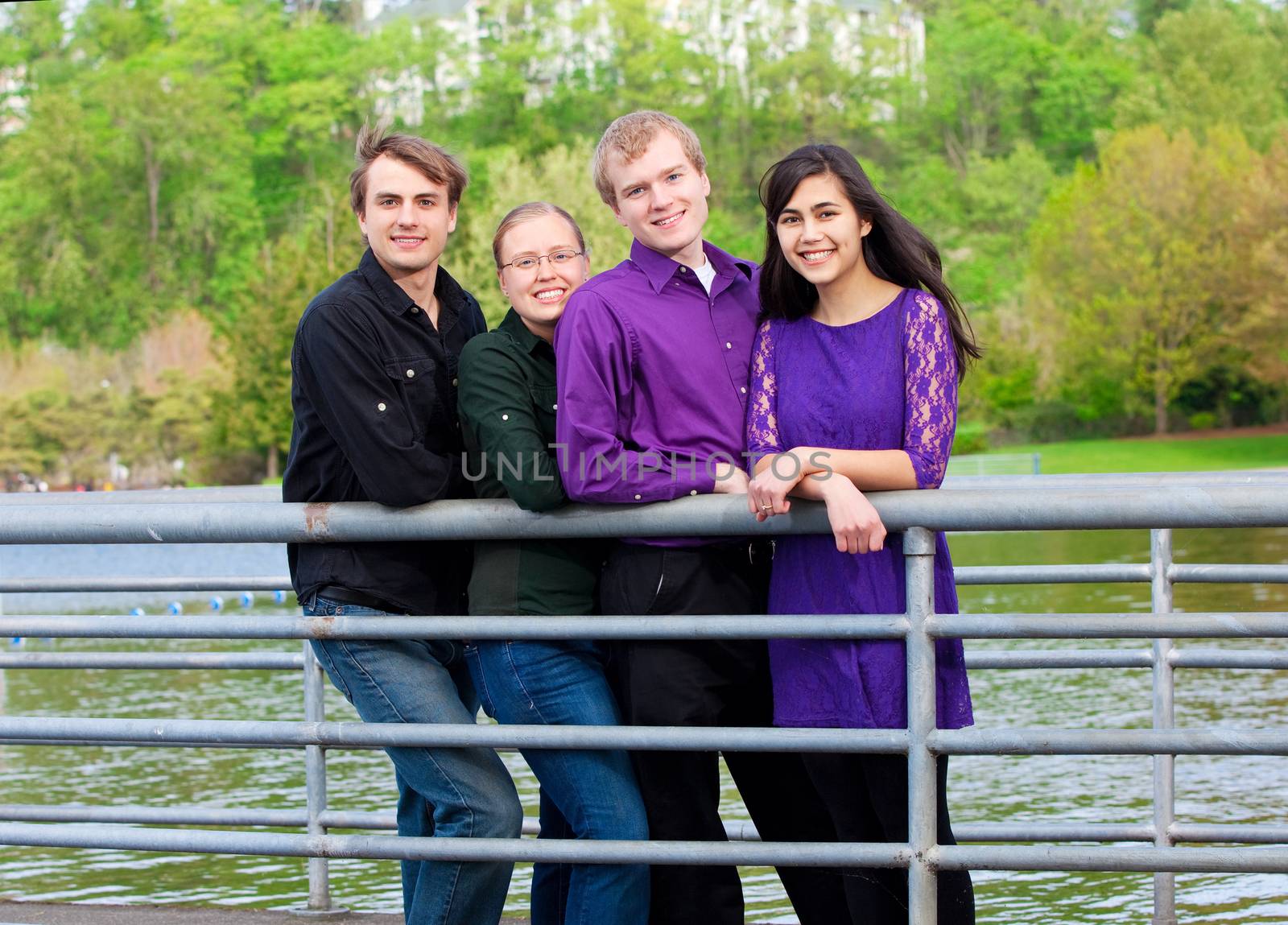 Four young multiethnic friends in early twenties standing  together along metal railing by lake