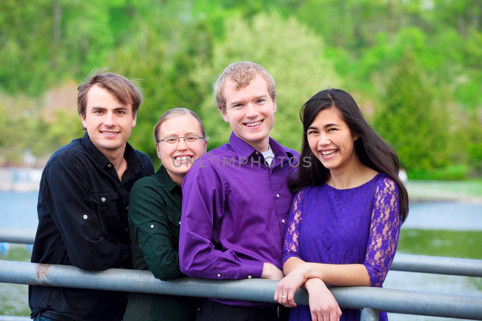 Four young multiethnic friends in early twenties standing  together along metal railing by lake