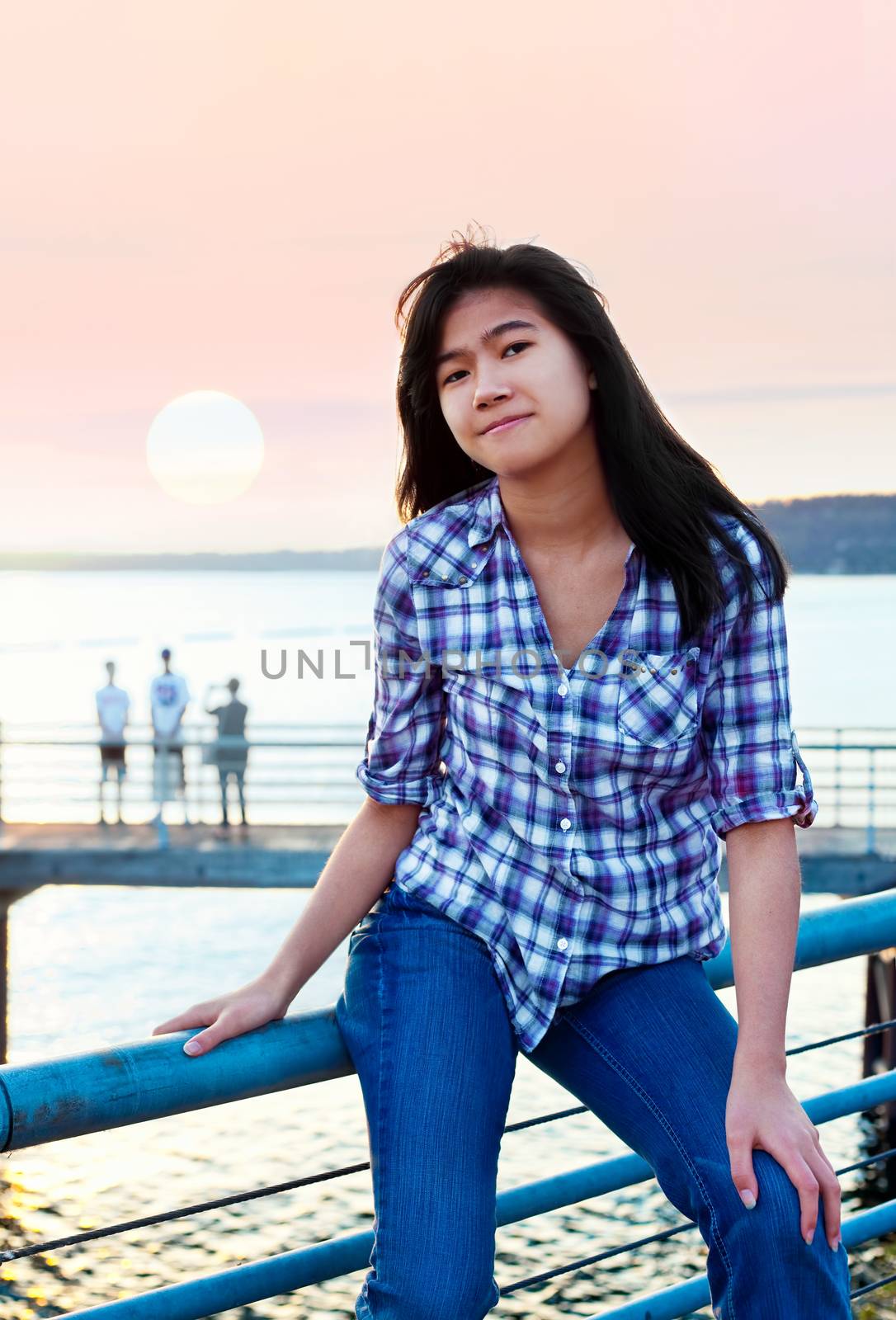 Young teen biracial Asian girl sitting on metal railing by lake at sunset, smiling and relaxing