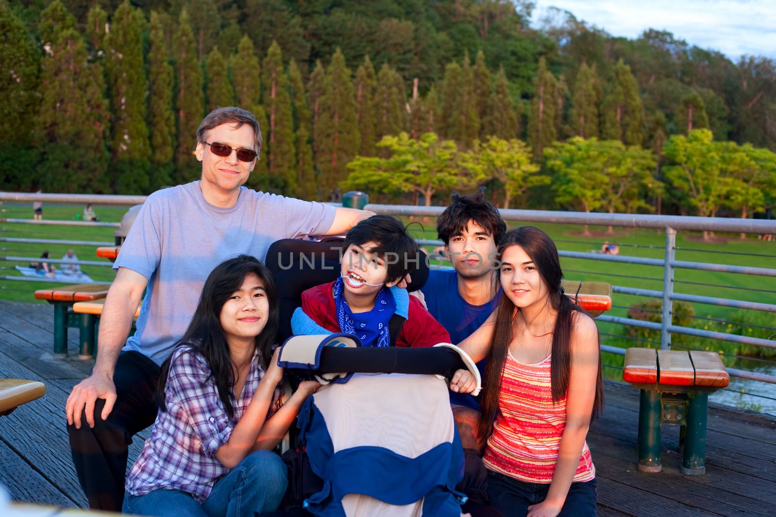 Caucasian father with biracial children at park, surrounding disabled son in wheelchair, Child has cerebral palsy.