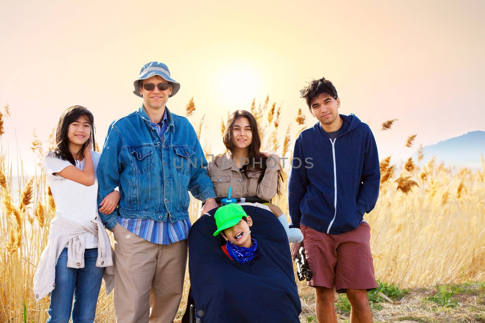 Caucasian father standing by field of tall grasses with biracial children. Youngest child in wheelchair has cerebral palsy.