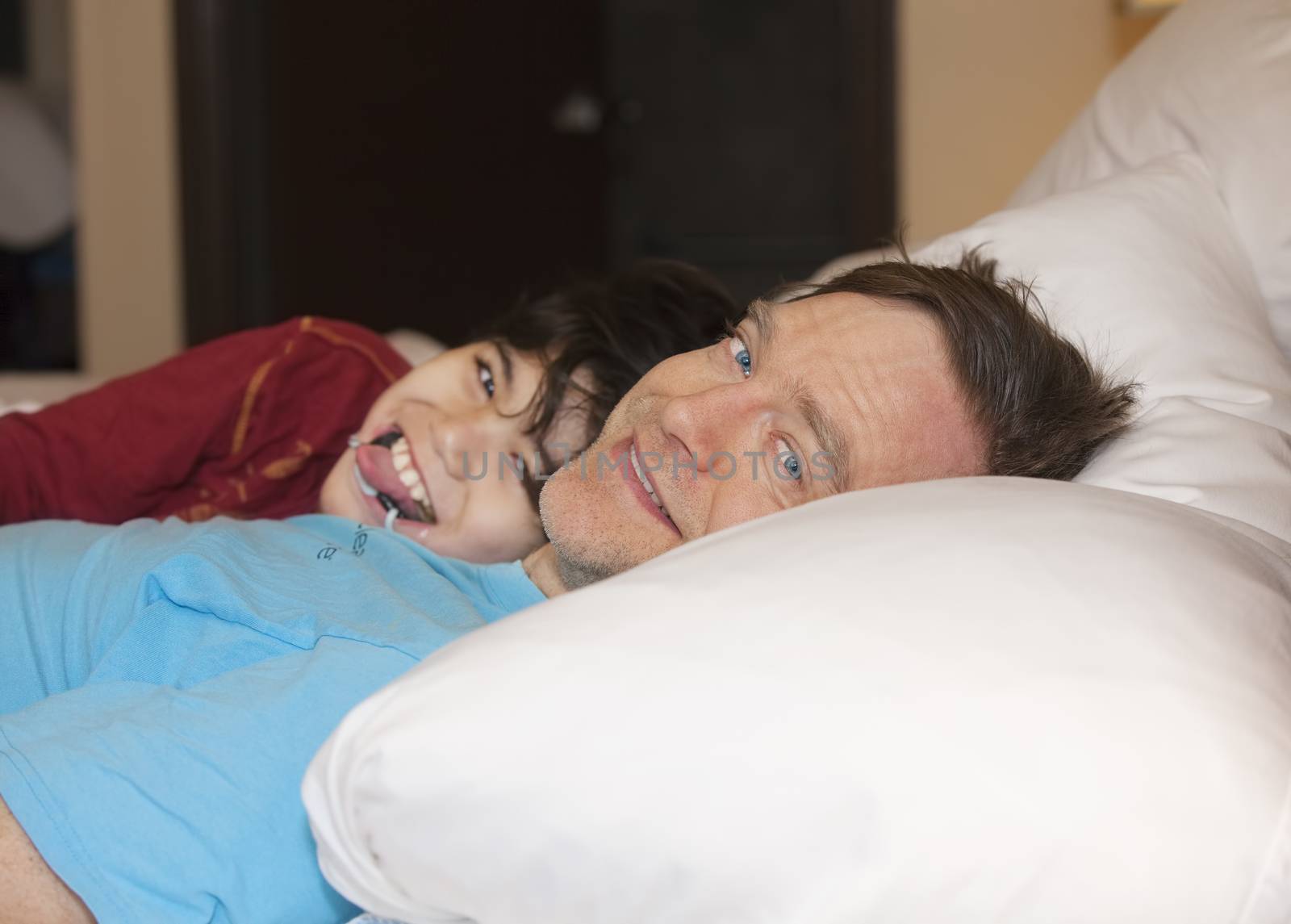 Caucasian father taking a nap with young disabled biracial son, in bed, laughing and talking together. Child has cerebral palsy. 