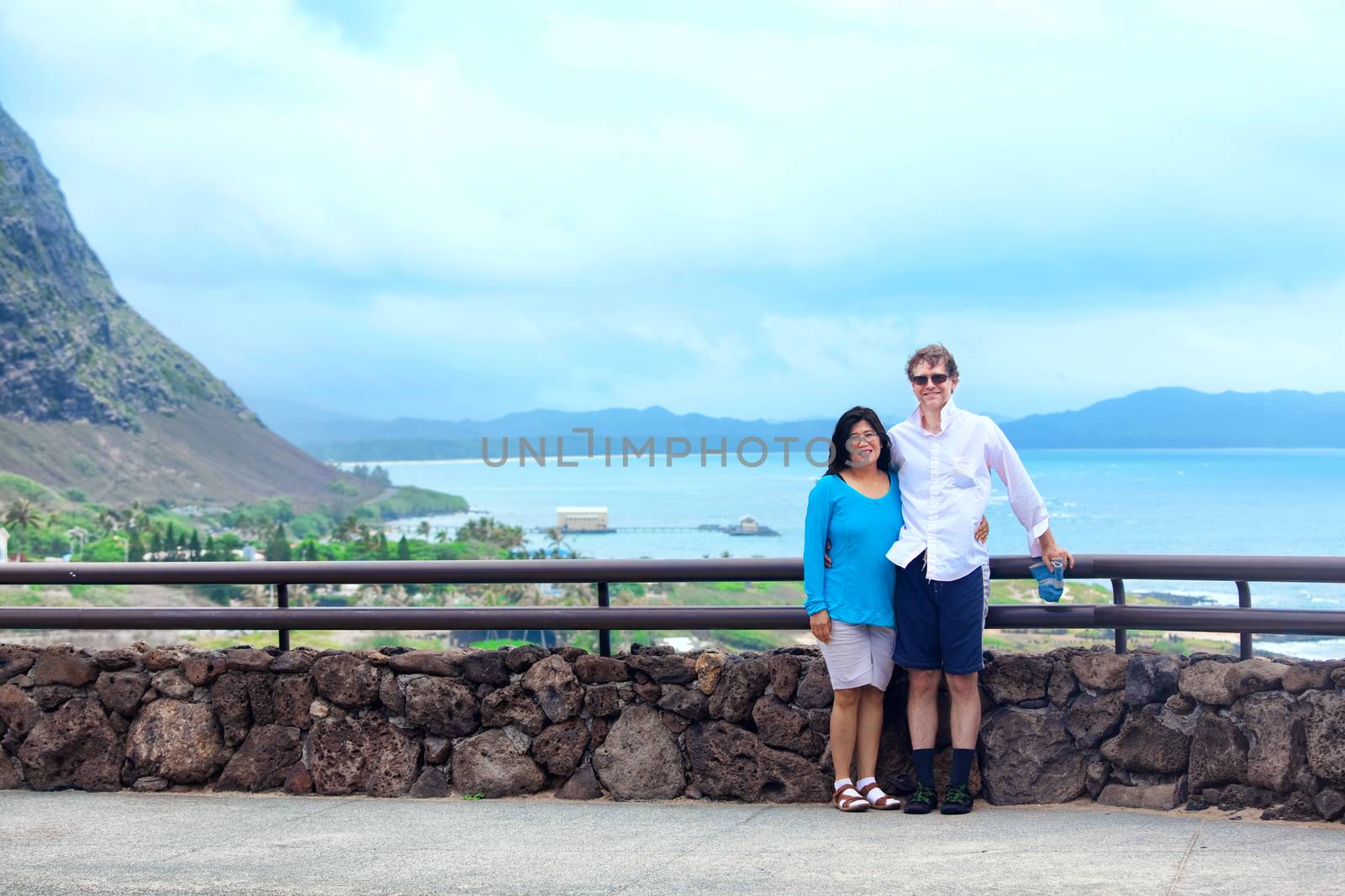 Interracial couple in forties standing by railing in Hawaii, with blue ocean scene in background