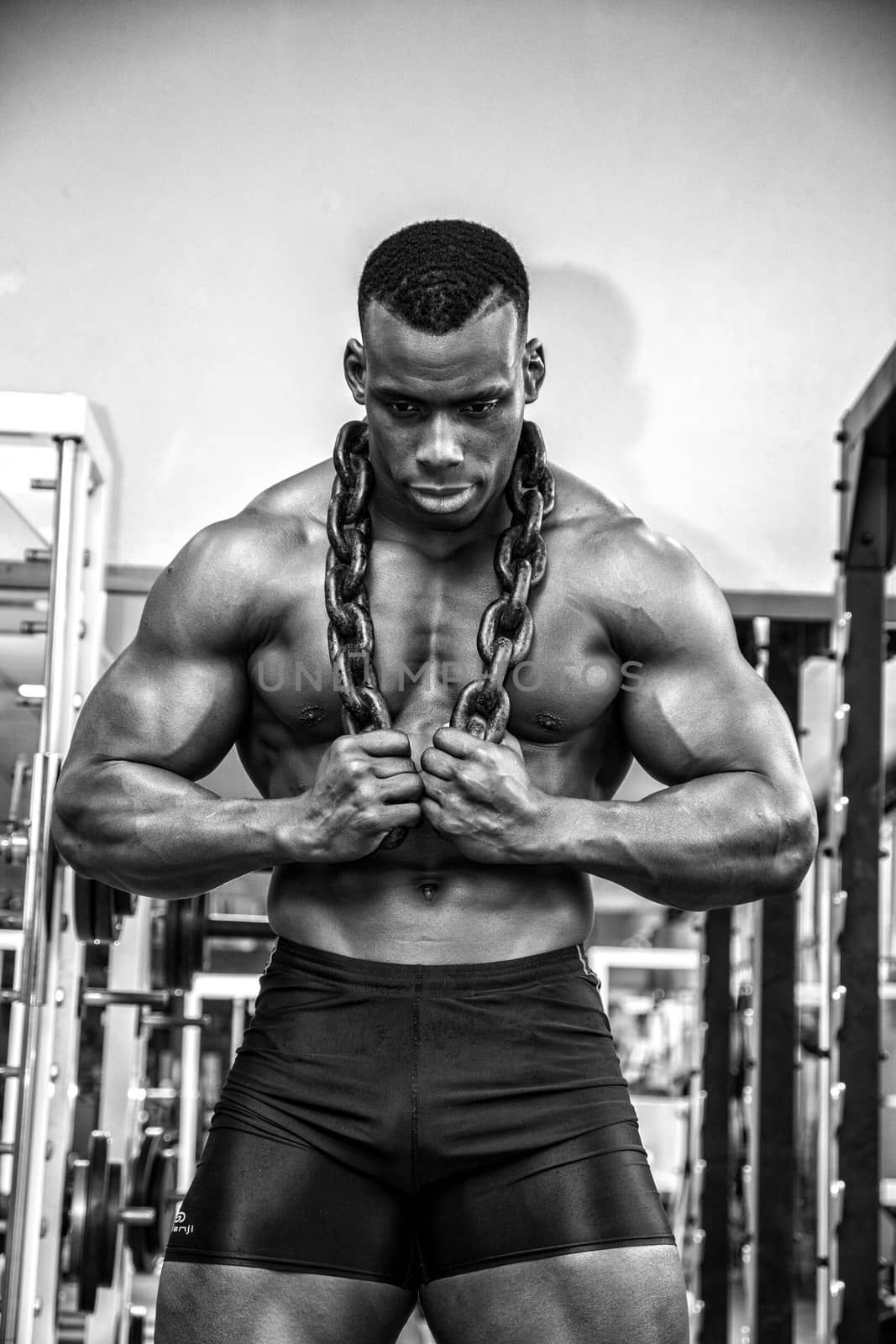 Attractive hunky black male bodybuilder doing bodybuilding pose in gym with iron chains over shoulders