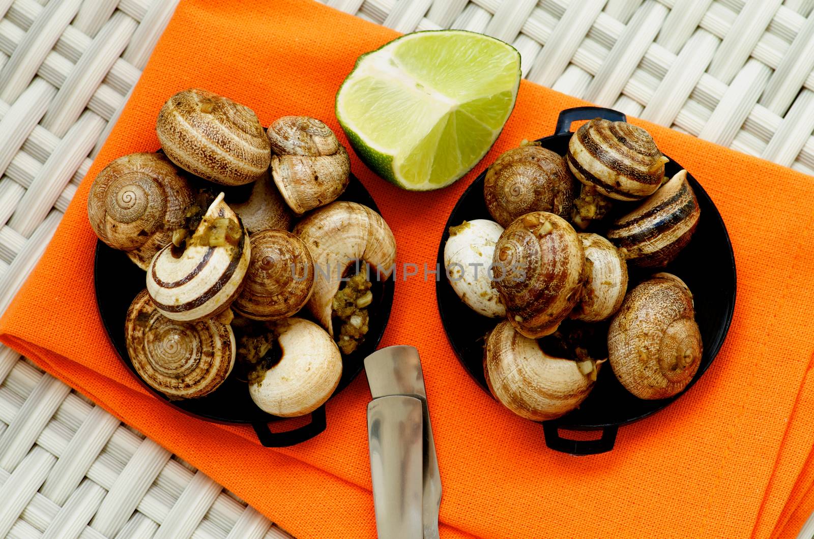 Delicious Escargot with Garlic Butter in Black Bowls with Sliced Lime closeup on Orange Napkin. Top View