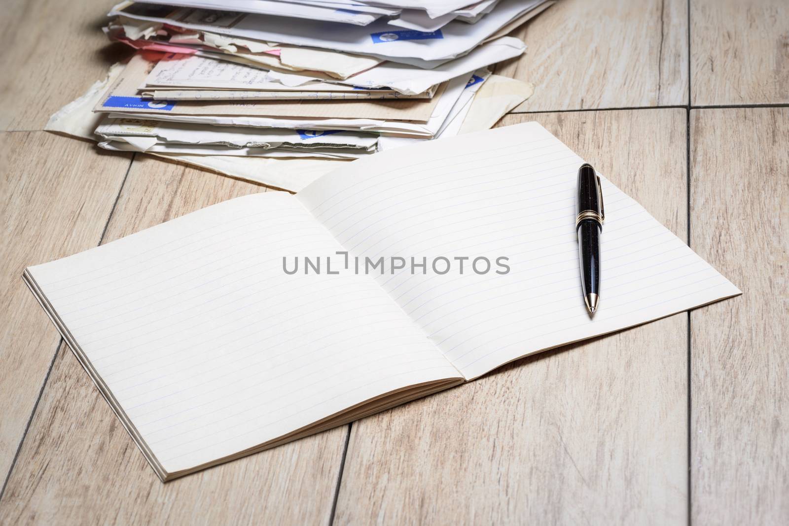 In the picture  a copybook with white sheets and a pen with letters in background.