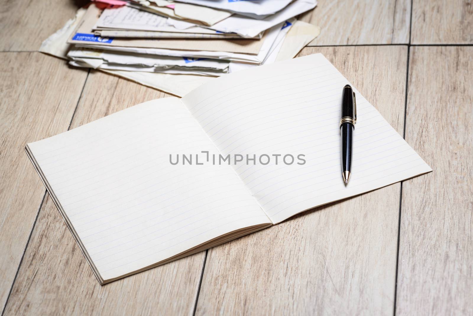 In the picture  a copybook with white sheets and a pen with letters in background.