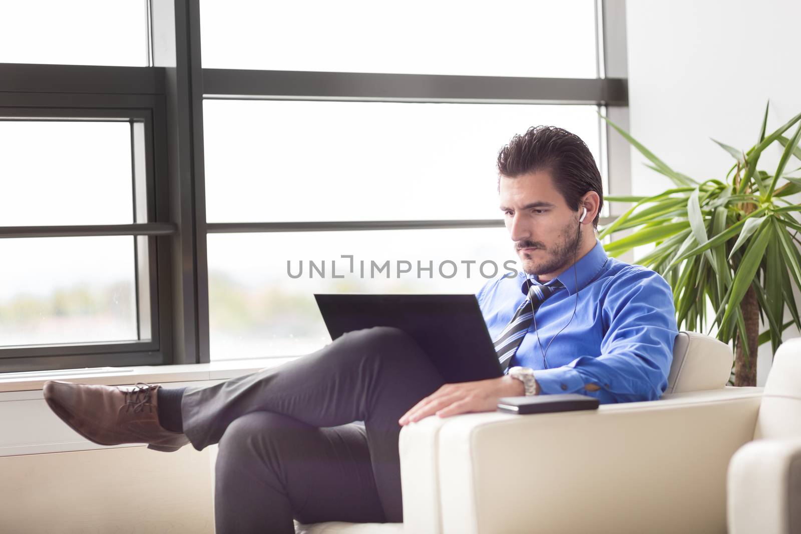 Portrait of successful young businessman in bright modern office focused on work on his laptop computer wearing headphones. Business and entrepreneurship concept.