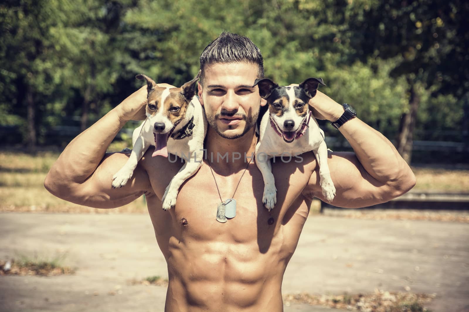 Half Body Shot of a Shirtless Athletic Young Man Carrying Two Dogs on his Shoulders and Looking at the Camera.