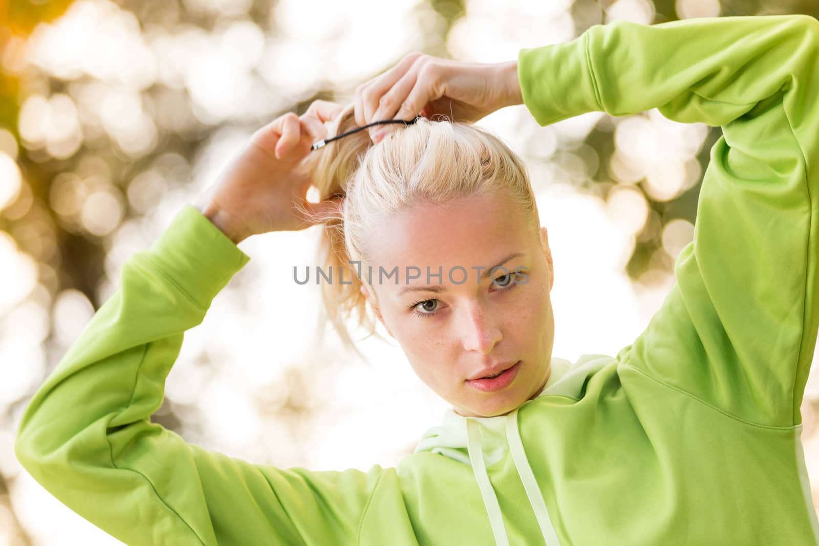 Portrait of attractive confident sporty woman wearing fashionable green sportswear  making her hair in ponytail before training in nature. Active sporty lifestyle concept.