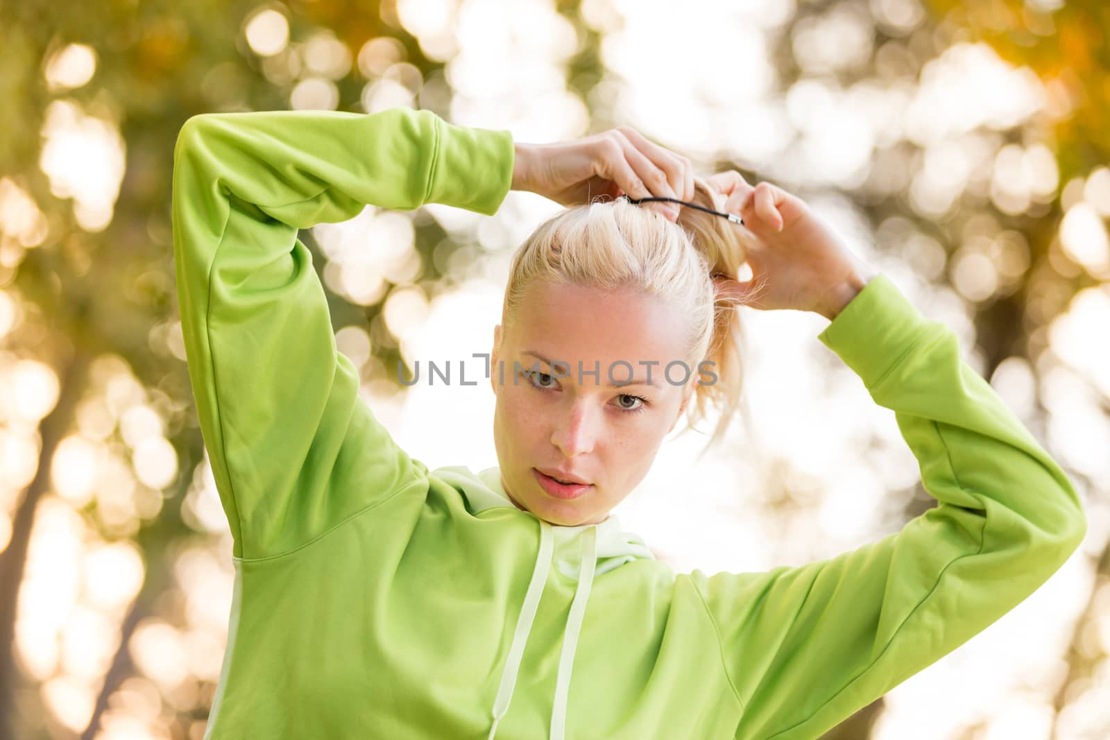 Portrait of attractive confident sporty woman wearing fashionable green sportswear  making her hair in ponytail before training in nature. Active sporty lifestyle concept.