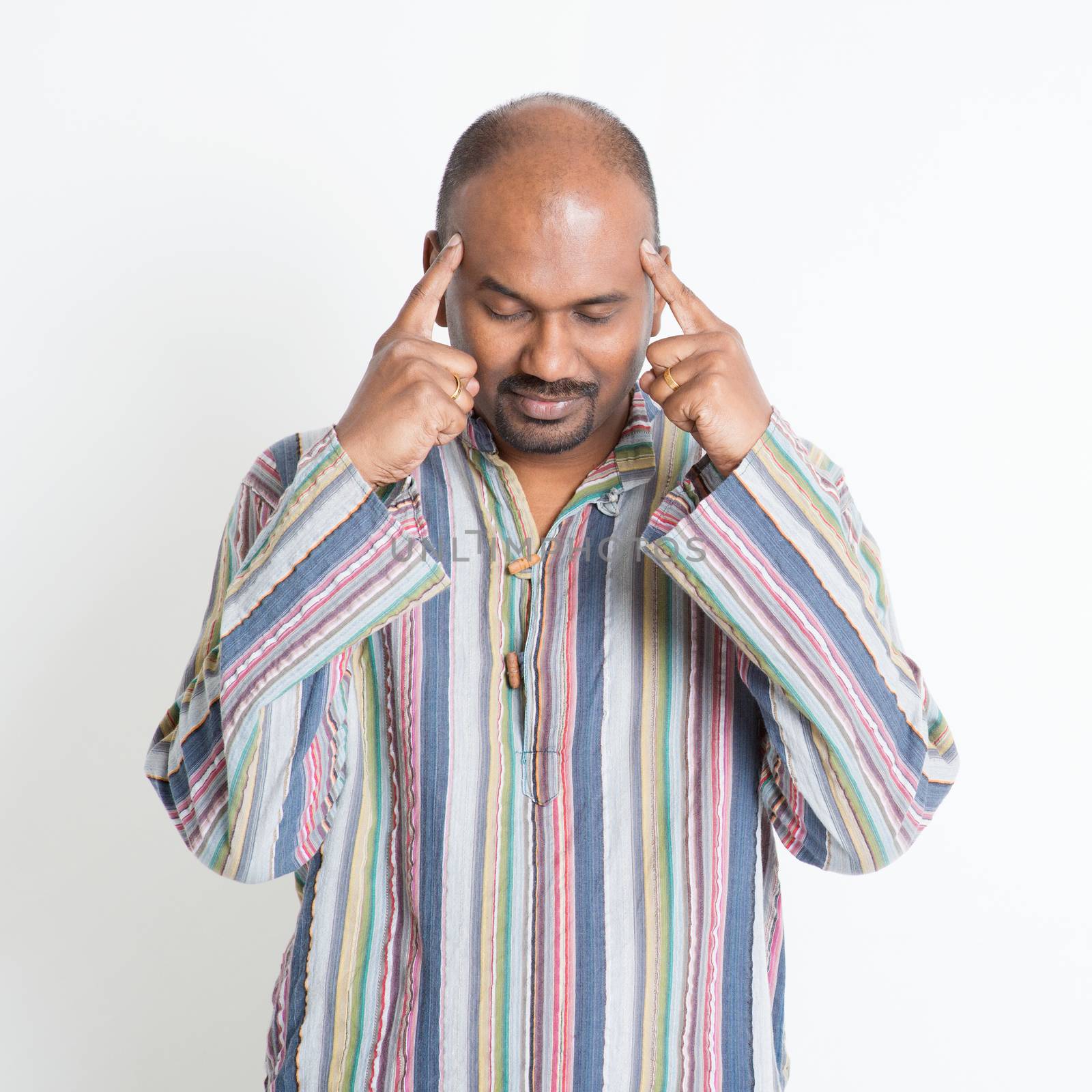 Portrait of mature casual business Indian man thinking, fingers pointing on head, standing on plain background.
