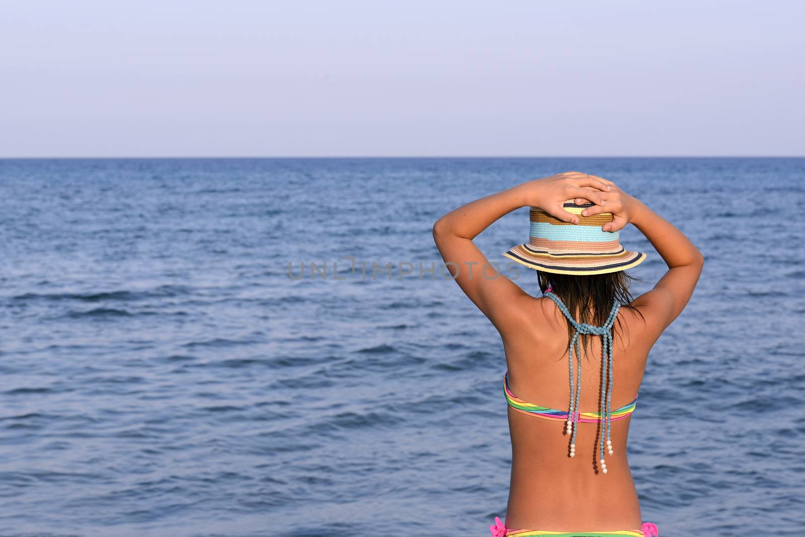 Little girl on the beach at sunset with her hands blocking a large hat.