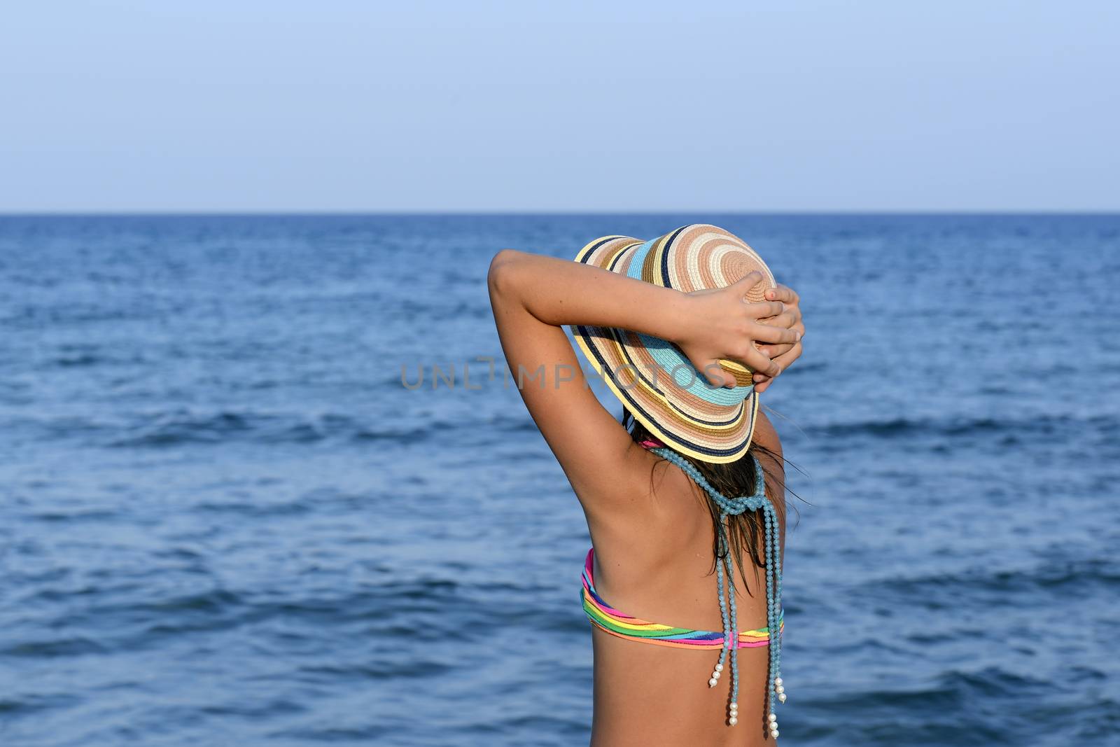 Little girl on the beach at sunset with her hands blocking a large hat.