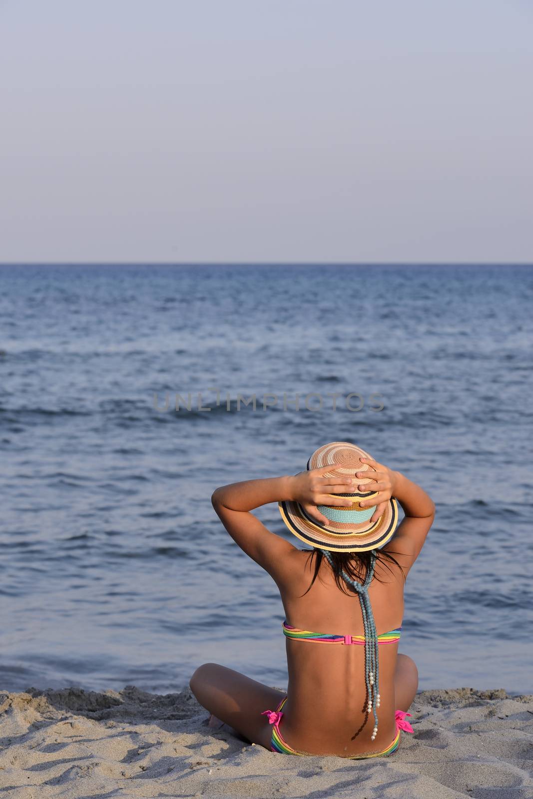 Little girl on the beach at sunset, with har hands blocking a large hat.