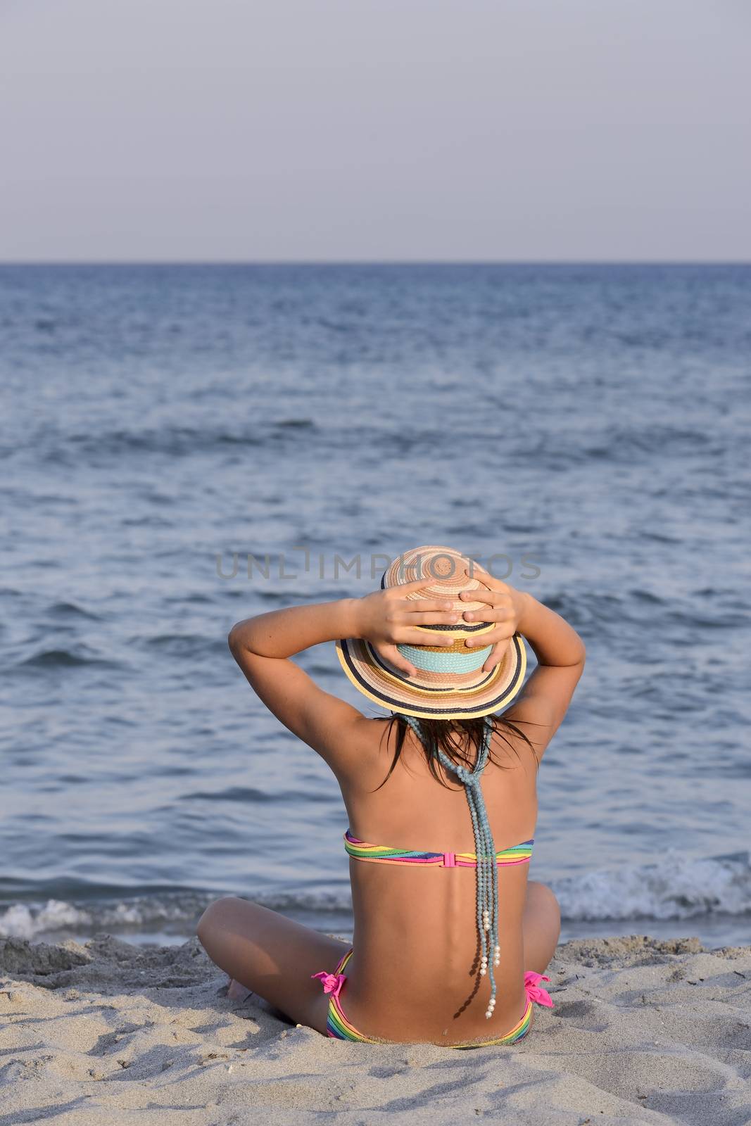 Little girl on the beach at sunset, with har hands blocking a large hat.