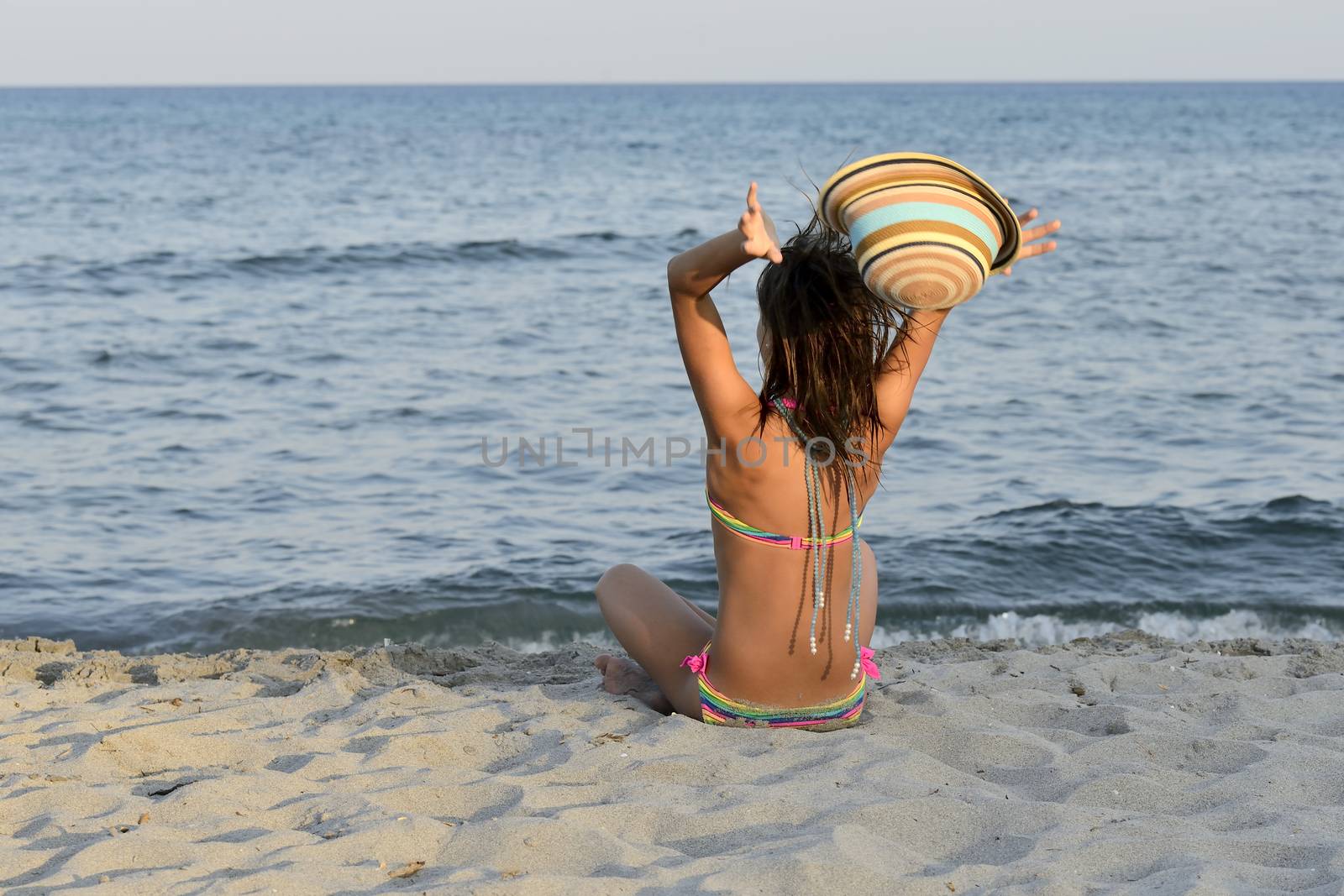 Little girl on the beach at sunset, with har hands blocking a large hat.