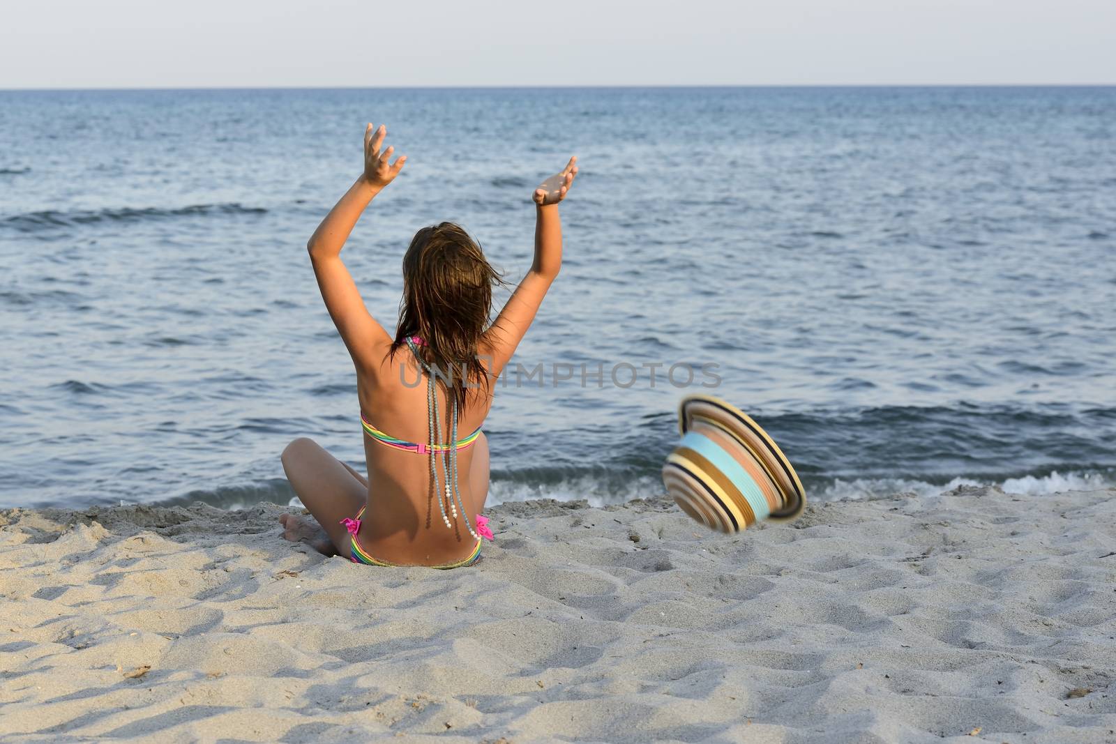 Little girl on the beach at sunset, with har hands blocking a large hat.