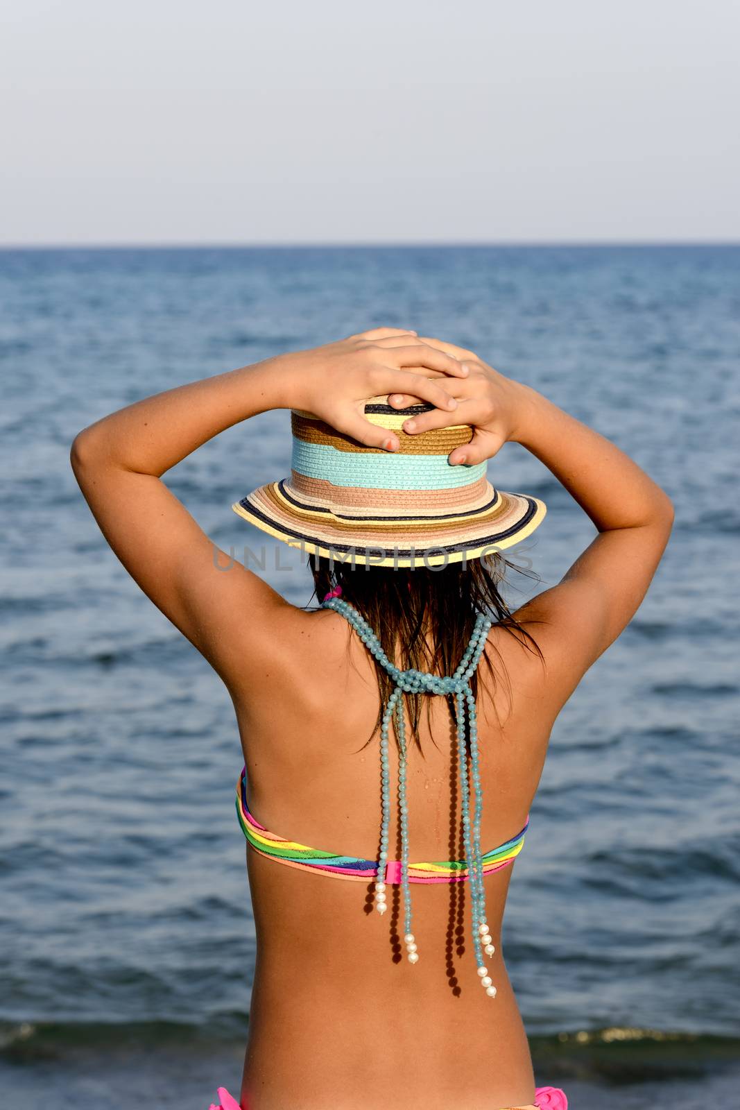 Little girl on the beach at sunset, with har hands blocking a large hat.