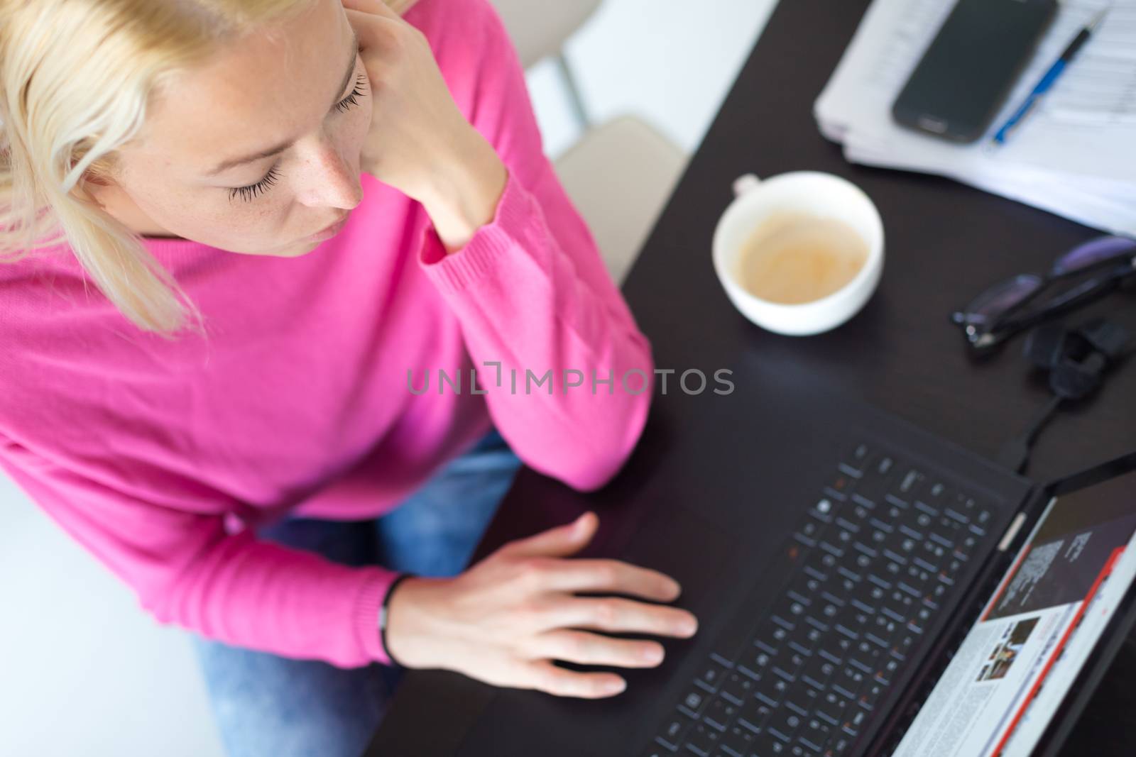 Female freelancer in her casual home clothing remotly working on laptop computer from her home while drinking her morning cup coffee. View from above.