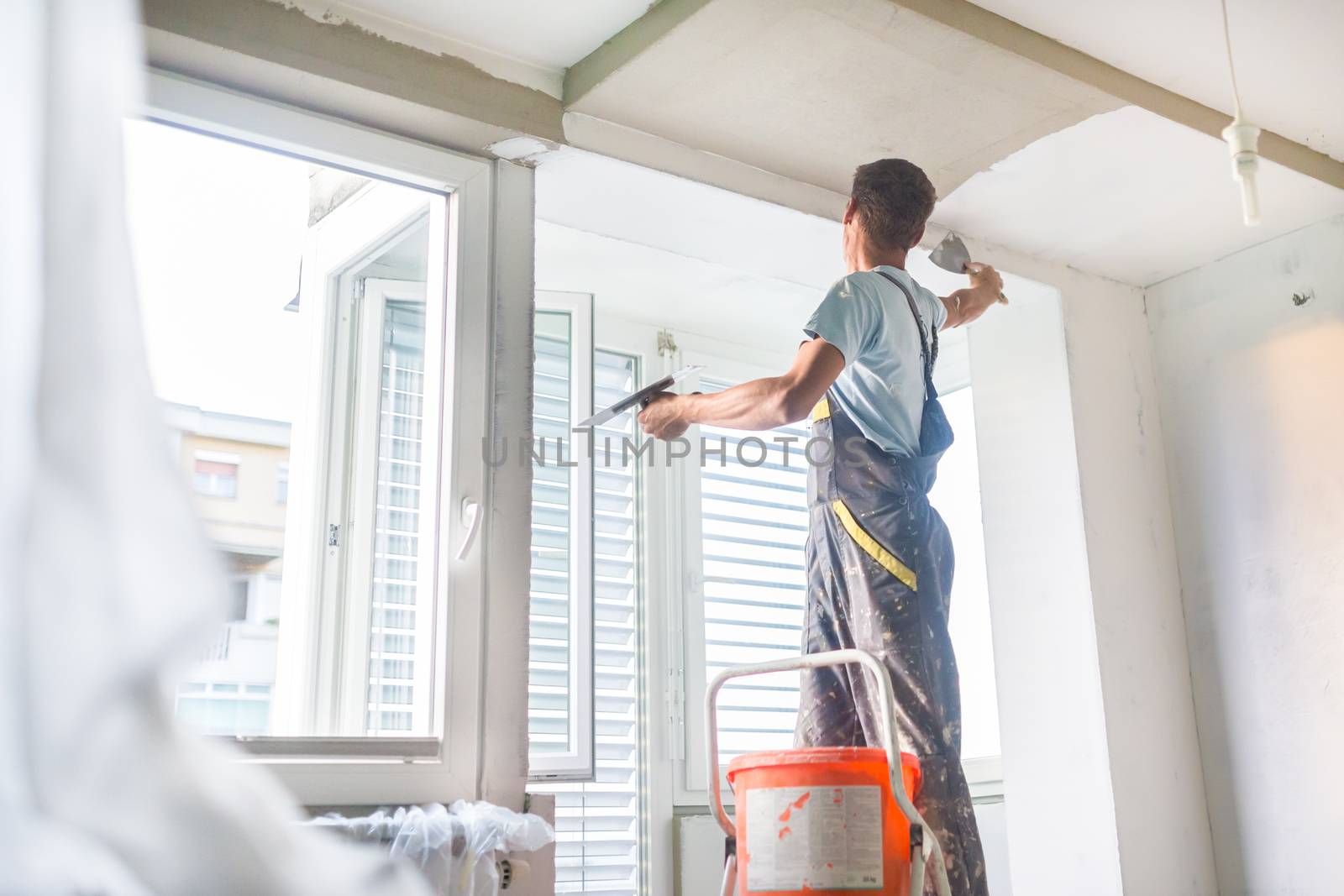 Thirty years old manual worker with wall plastering tools inside a house. Plasterer renovating indoor walls and ceilings with float and plaster.